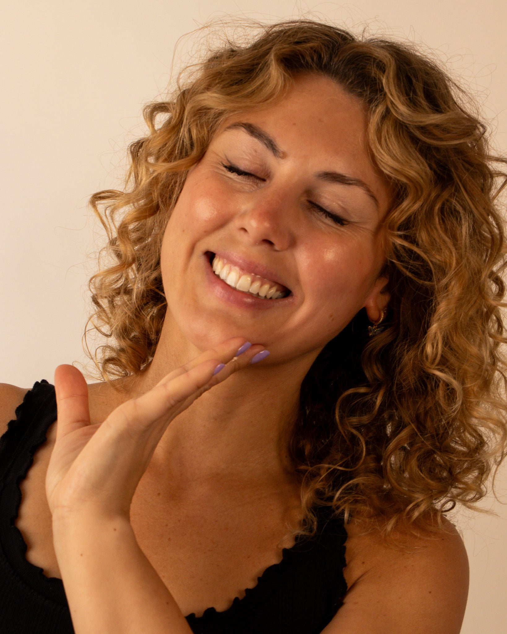 Woman with curly hair smiling and touching her face against a plain background