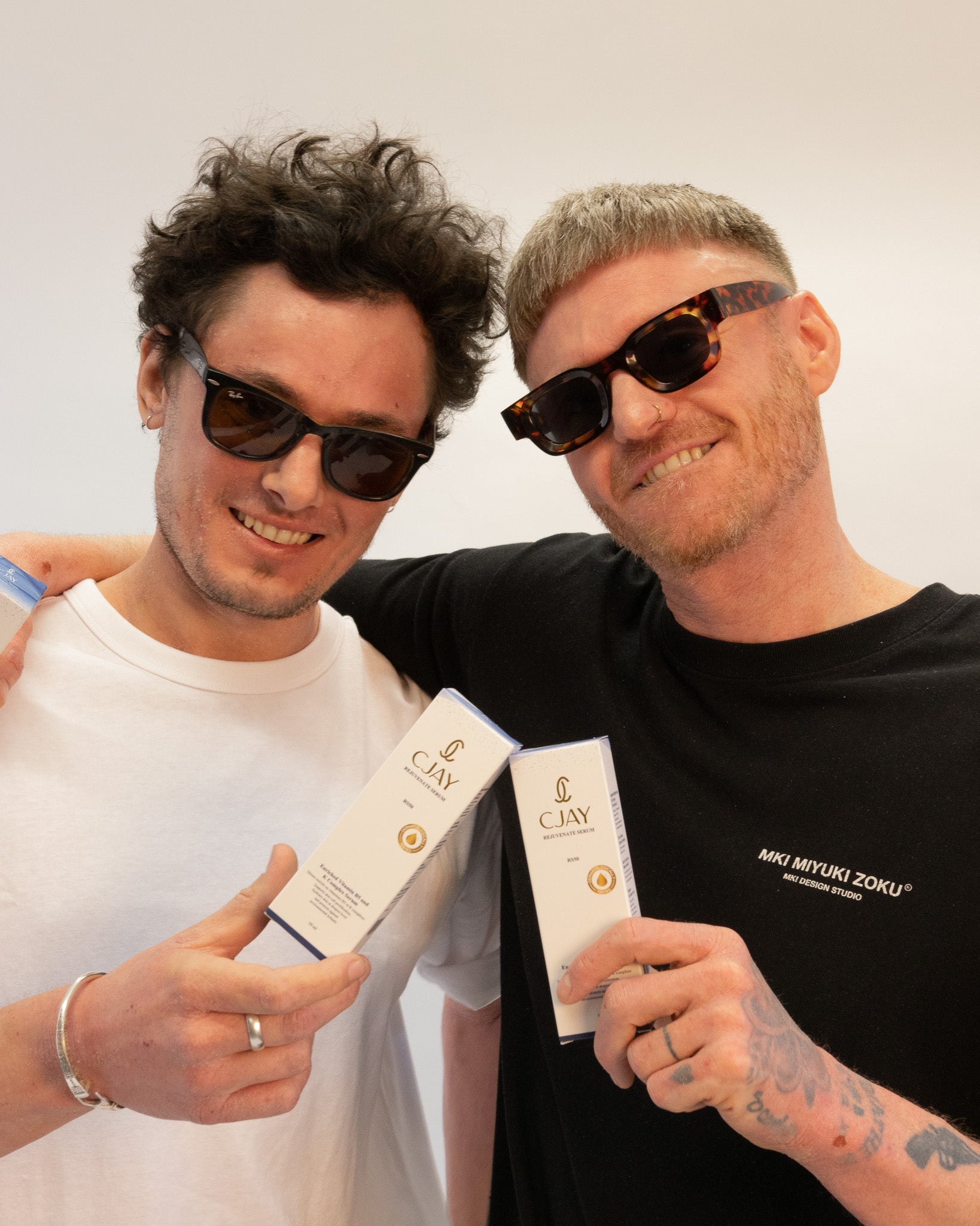 Two men wearing sunglasses and holding CJAY products against a plain background